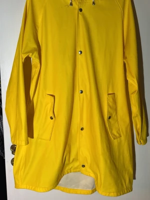 Gul regnjacka från 157 med huva - Klassisk gul regnjacka från 157 i storlek XL. Jackan har huva med vit dragsko, tryckknappar framtill och två fickor med lock. Tillverkad i syntetmaterial som står emot regn, perfekt för blöta dagar. Enkel och stilren design.