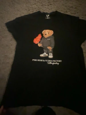 Svart t-shirt Pyro Bear Sector Ultras - Svart t-shirt från Sector Ultras med tryck av en björn i hoodie som håller en fackla. Texten 'PYRO BEAR by ULTRAS FACTORY' under motivet. Klassisk passform och rund hals. Perfekt för dig som gillar streetwear och coola prints.