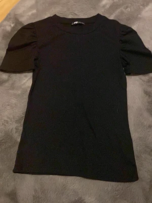 Svart topp med puffärm - Snygg svart topp från FB Sister i ribbadt bomull material med puffiga korta ärmar. Enkel rund halsringning och tight passform som ger en clean look. Perfekt att styla med jeans eller kjol för en trendig outfit.