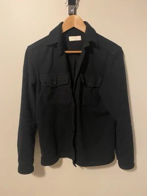 Svart overshirt från Jack n Jones  - Svart overshirt från Schnayderman's i bomull med klassisk krage, två bröstfickor med knappar och raka ärmar. Enkel och stilren design som passar perfekt till jeans eller chinos. Långärmad och normal passform.