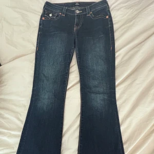 True Religion mörkblå bootcut jeans - Snygga mörkblå jeans från True Religion med klassisk bootcut och kontrastsömmar. Jeansen har ikonisk fickdesign med lock och knapp bak, samt detaljerade sömmar fram. Perfekt passform och cool look för dig som vill sticka ut.