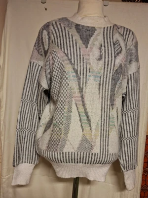 Mönstrad stickad tröja från 18 Carat - Unik stickad tröja från 18 Carat med coolt grafiskt mönster i grått, vitt och pastellfärger. Tröjan har långärmade ribbade muddar och rund hals. Mönstret är en mix av ränder och geometriska former som ger en retro vibe. Perfekt för dig som gillar statement-plagg.