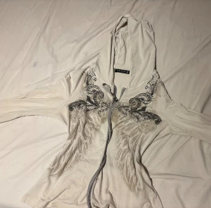 Y2K hoodie från Betty Barclay - As snygg Y2K hoodie med ving och svarta diamant detaljer! Säljer pga de inte är min stil längre. Tyvärr så har den är hål rakt vid blixtlåset, fast de syns knappt om man har nåt vitt under!  Typ mellan en S-M 