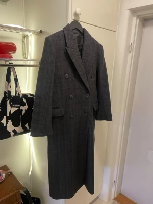 Mörkgrå rutig lång kappa ull Weekday  - Snygg lång kappa i mörkgrått med diskret rutigt mönster och dubbelknäppning framtill. Jackan har breda slag, två fickor med lock och är helfodrad. Perfekt för dig som vill ha en klassisk och stilren look under höst och vinter. Lite för liten för mig så därför får den gå vidare till någon annan. Endast använt den ett par gånger och därmed är den i nästintill nyskick