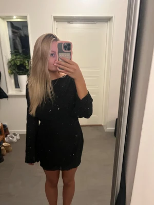 Svart glittrig kortklänning med öppen rygg - Säljer en svart kortklänning med glittriga detaljer och lång ärm. Klänningen har en snygg öppen rygg med knytband upptill. Är som en small i storlek. Pris kan diskuteras!💕