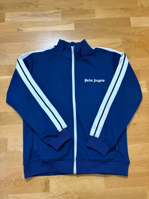 Blå Palm Angels zip hoodie med vita ränder - Snygg marinblå zip hoodie från Palm Angels med vita och svarta ränder längs ärmarna. Tröjan har hög krage, lång ärm och logotypen broderad på bröstet. Tillverkad i mjuk polyester med ribbade muddar och hel dragkedja framtill.