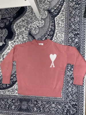 Rosa stickad tröja från Ami Paris - Snygg rosa stickad tröja från Ami Paris med ett vitt hjärta och bokstaven A på bröstet. Tröjan har rund hals, ribbade muddar och långärmad passform. Perfekt för dig som gillar stilrena detaljer och vill ha något som sticker ut lite extra.Denna är unisex 