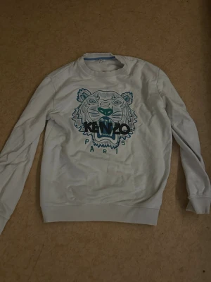 Vit Kenzo sweatshirt med tigertryck - Säljer en vit sweatshirt från Kenzo med det ikoniska tigertrycket och Kenzo Paris-loggan i blått och turkost framtill. Tröjan har rund hals, långa ärmar och ribbade muddar. Perfekt för dig som vill sticka ut med en klassisk streetstyle-look.