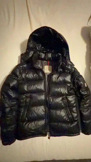 Svart Moncler pufferjacka med huva - Svart pufferjacka från Moncler med glansig finish, huva och dragkedja framtill. Jackan har Moncler-logga på ärmen och praktiska fickor med dragkedja. Perfekt för kalla dagar.