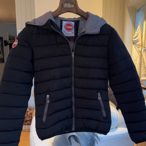 Svart pufferjacka från Colmar - Svart pufferjacka från Colmar Originals med huva och grått foder. Jackan har två sidofickor med dragkedja och en snygg logga på ärmen. Perfekt för kyliga dagar och har en stilren, quiltad design med raka linjer.