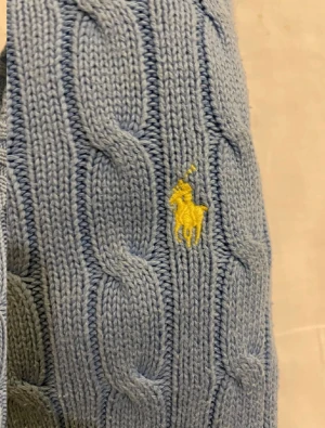 Ljusblå stickad tröja Polo Ralph Lauren - Snygg ljusblå stickad tröja från Polo Ralph Lauren med klassiskt kabelstickat mönster och gul broderad logga på bröstet. Rund halsringning och ribbade muddar vid ärmslut och nederkant. Perfekt för dig som gillar stilrena och tidlösa plagg.