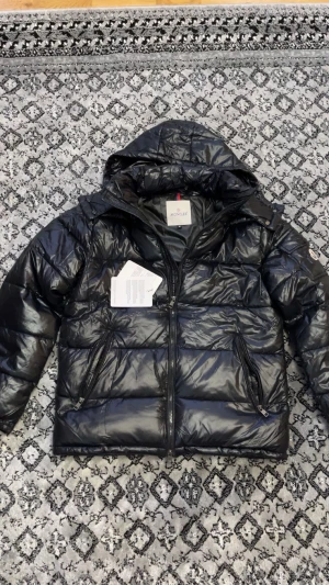 Svart Moncler pufferjacka med huva - Svart pufferjacka från Moncler med glansig finish, huva och flera dragkedjefickor. Jackan har klassisk Moncler-logga på ärmen och coolt serietryck på insidan. Perfekt för dig som vill ha en varm och snygg jacka med streetkänsla.