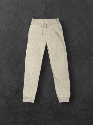 Grå mjukisbyxor från Real Performance - Snygga grå mjukisbyxor från Real Performance med ribbade muddar och snörning i midjan. Byxorna har två sidofickor och en mjuk, skön känsla tack vare bomullsmaterialet. Perfekta för chill eller träning, med stilren look och bekväm passform.