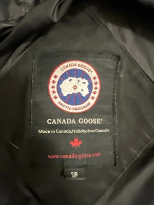 Svart Canada Goose dunjacka med päls - Svart lång dunjacka från Canada Goose med ikonisk röd och vit patch på ärmen. Jackan har huva med beige päls, dragkedja och flera fickor. Perfekt för kalla vinterdagar och har en stilren look som funkar till allt. Klassisk modell med snygga detaljer och varm fyllning.