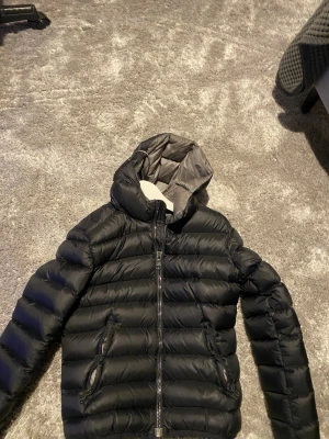 Svart dunjacka från Colmar med huva - Säljer en svart dunjacka från Colmar med huva och två sidofickor med dragkedja. Jackan har ett stilrent, quiltat mönster och logotyp på ärmen. Perfekt för kalla dagar och har en snygg, modern look som funkar till många stilar.