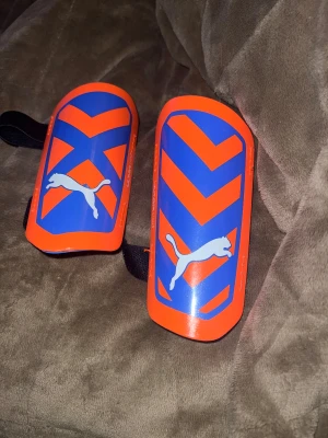 Blå och orange benskydd från Puma - Säljer ett par snygga benskydd från Puma i blått och orange med coola grafiska mönster och vit Puma-logga. De är ultralätta och har svarta elastiska band för att sitta på plats under matchen. Perfekta för dig som vill sticka ut på fotbollsplanen. För barn