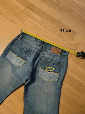 Diesel Larkee blå jeans straight fit - Säljer ett par klassiska Diesel Larkee jeans i blå tvätt med snygga slitningar och kontrastdetaljer på bakfickorna. Modellen har rak passform, normal midja och är tillverkad i mjukt jeanstyg med gul söm. Perfekt för dig som gillar en chill och tidlös look.