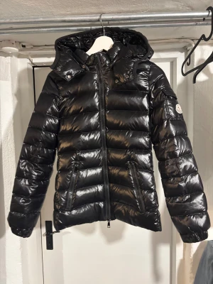  Moncler Bady jacka - Svart pufferjacka från Moncler med glansig finish, huva och två dragkedjeförsedda fickor. Jackan har Moncler-logga på ärmen och serietidningsfoder på insidan. Perfekt för dig som vill ha en snygg och varm jacka. Skriv privat för fler bilder eller frågor😊 har nfc tag och påsen. Storlek 1 vilket motsvarar S, men passar xs o m också