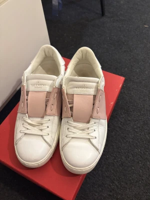 Valentino Garavani skor  - Snygga Valentino Garavani sneakers i vitt skinn med ljusrosa detaljer och klassiska nitar baktill. Skorna har rund tå, platt sula och snörning framtill. Perfekt för dig som gillar lyxiga och trendiga sneakers med unik design. Väldigt fina i sitt skick ser som nya ut, sulan är väldigt bra också knappt slitet.