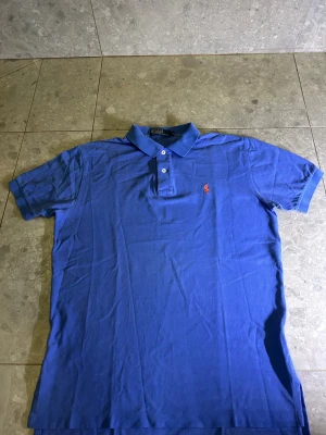 Blå Polo Ralph Lauren pikétröja M - Snygg blå pikétröja från Polo Ralph Lauren i klassisk pikémodell med krage och två vita knappar.  Skulle säga att den är lite urtvättat! Pris är diskuterbart vid snabb affär!
