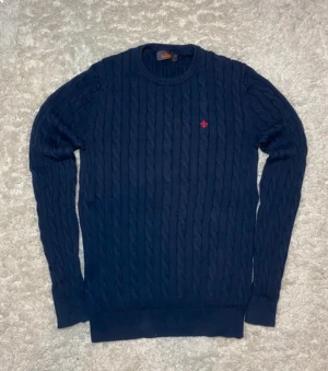 Marinblå kabelstickad tröja Morris - Säljer en marinblå kabelstickad tröja från Morris med rund hals och diskret röd logga på bröstet. Tröjan är gjord i mjuk pima bomull och har långa ärmar samt ribbade muddar. Perfekt för dig som gillar klassisk stil med snygga detaljer.