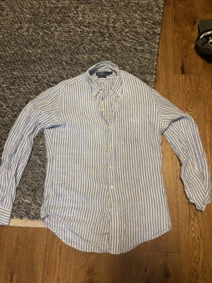 Blåvit randig skjorta från Polo Ralph Lauren - Snygg blåvit randig skjorta från Polo Ralph Lauren i storlek S. Skjortan har klassisk krage med knappar, lång ärm och en relaxed passform. Tillverkad i mjukt linne, perfekt för en chill och stilren look. Custom Fit-modell med knappar framtill.