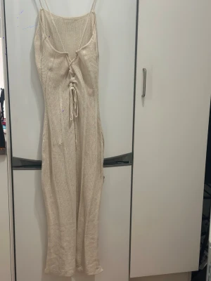 Beige stickad långklänning med snörning L - Snygg beige stickad långklänning med smala axelband och snörning framtill. Klänningen har en lätt transparent känsla och slits på sidan för extra vibe. Perfekt för sommarens varma dagar och har en loose passform som ger en chill look.