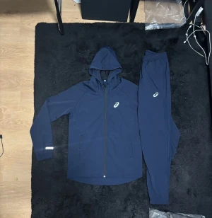 Marinblå Asics träningsset finns i alla storlek  - Snyggt marinblått träningsset från Asics med matchande hoodie och byxor. Setet har dragkedja på tröjan, huva och Asics-logga på både överdel och underdel. Perfekt för dig som gillar sportig stil och vill ha något bekvämt till gymmet eller löprundan.