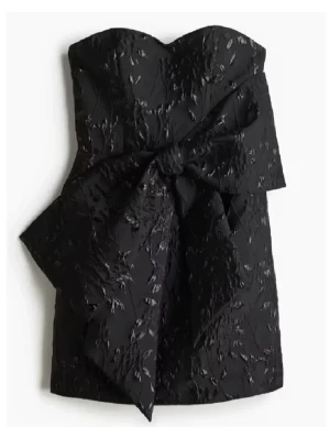 Svart axelbandslös kort klänning med rosett - Snygg svart axelbandslös kort klänning med strukturerat tyg och en stor rosett framtill. Klänningen har en elegant och modern look med en figurnära passform och subtil glans i materialet. Perfekt för dig som vill sticka ut. 