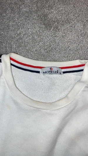 Vit långärmad Moncler tröja XL - Vit långärmad tröja från Moncler i storlek XL. Snygga svarta band med Moncler-logga längs båda ärmarna och en krage med röd, vit och blå detalj. Tröjan har en klassisk passform och är tillverkad i mjuk bomull som känns skön mot huden.