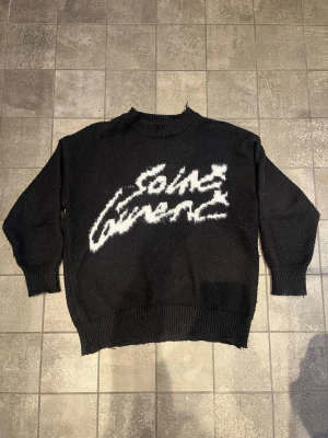 Saint Laurent svart stickad tröja - Snygg svart stickad tröja från Saint Laurent med vit, fluffig logotyp framtill. Tröjan har ribbade muddar vid hals, ärmslut och nederkant samt långärmad passform. Perfekt för dig som gillar exklusiva och trendiga plagg med statement-detaljer.