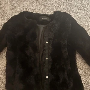 Svart pälsjacka från Vero Moda - Säljer en svart pälsjacka från Vero Moda med rund hals och knappar framtill. Jackan har en fluffig och mjuk yta som ger en lyxig vibe. Perfekt för dig som vill ha en snygg och varm jacka med stilren look.