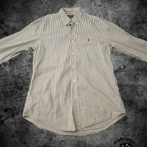 Randig skjorta Polo Ralph Lauren M - Snygg randig skjorta från Polo Ralph Lauren i slim fit. Skjortan är vit med mörkblå ränder, har klassisk krage och knappar framtill. På bröstet finns det ikoniska röda Polo-logot. Perfekt för dig som gillar stilrena och tidlösa plagg, den är fläckfri och har noll defekter. Skriv om det finns några funderingar, pris kan diskuteras vid snabb affär!