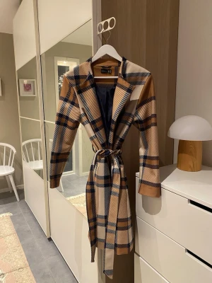 Rutig kappa med bälte  - Snygg rutig kappa i beige, brunt och svart med klassisk krage och bälte i midjan. Långärmad och med normal passform, perfekt för dig som gillar statement-plagg med stilren vibe. Materialet är mjukt och känns lyxigt, och kappan har ett helfodrat svart innerfoder. Storlek 36.