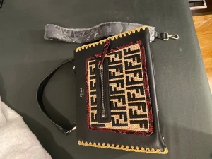  svart axelväska med mönster - Svart axelväska från skinn med ikoniskt mönster i beige och svart på framsidan, omringat av röda detaljer. Väskan har gul dekorativ kant, dragkedja och avtagbar axelrem. Perfekt storlek för det viktigaste och har en cool, lyxig vibe.