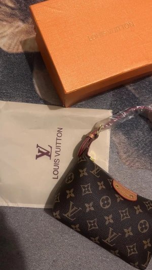 Louis Vuitton monogram axelväska - Snygg axelväska från Louis Vuitton med klassiskt monogrammönster i brunt och beige. Väskan har guldfärgade detaljer, dragkedja och en stilren form. Insidan har randigt foder och en logotyp i läder. Perfekt för dig som vill ha en ikonisk designeraccessoar.