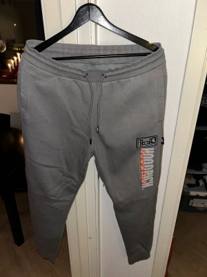 Grå Hoodrich mjukisbyxor med tryck - Snygga grå mjukisbyxor från Hoodrich med coolt tryck och broderad logga i vitt och orange på benet. Byxorna har snörning i midjan, ribbade muddar och detaljer i orange. Perfekta för streetwear och chill dagar.