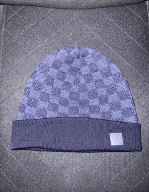 Blå rutig mössa från Louis Vuitton - Snygg blå mössa från Louis Vuitton med klassiskt rutigt mönster och ribbad kant. Märkets logga i blått läder sitter framtill. Mössan är stickad och har en modern, urban vibe som passar perfekt till streetwear.