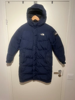  dunjacka The North Face - Säljer en mörkblå lång dunjacka från The North Face i storlek 150. Jackan passar för barn som är ungefär 146–151 cm långa. Den har huva, dragkedjeförsedda fickor och en quiltad design med logga på bröstet och ärmen. Perfekt för kalla dagar och håller barnet varmt och bekvämt.  Pris kan diskuteras. Skicks gärna, bud.