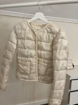 Beige quiltad pufferjacka med flätad kant - Snygg beige pufferjacka med quiltad design och rund hals. Jackan har flätad kant vid hals och fickor, samt två framfickor och knappar framtill. Perfekt för dig som gillar stilrena och trendiga jackor med unika detaljer.