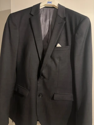 Mörkgrå kostym från Matinique, strl 48 - Säljer en stilren mörkgrå kostym från Matinique i storlek 48. Kavajen har klassisk passform med två knappar, ficklock och bröstficka med vit näsduk. Byxorna är raka och matchar kavajen perfekt. Materialet känns mjukt och har en lätt glans.