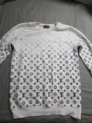 Vit Louis Vuitton sweatshirt S - Vit sweatshirt från Louis Vuitton i storlek S med ikoniskt svart LV-mönster över hela tröjan. Rund hals och långa ärmar, tillverkad i mjuk bomull. Perfekt för dig som gillar lyxiga streetwear-vibbar och vill sticka ut med ett klassiskt designertryck. Har ett litet hål vid vänster armbåge.