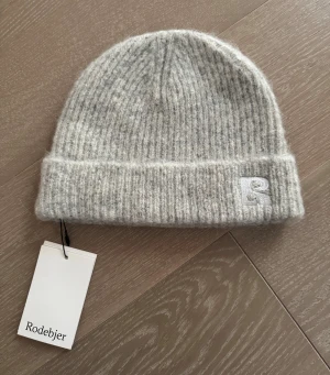 Rodebjer ny grå stickad mössa alpacka ull helt ny - Mössan är gjord av ett mjukt och varmt material, perfekt för att hålla huvudet varmt under de kallare månaderna. Beanien har en enkel design med en liten logotyp på framsidan. 57% alpacka  25% ull  18% nylon Helt ny