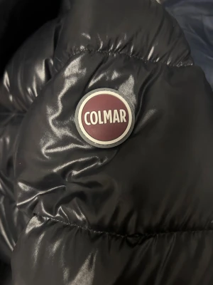 Svart pufferjacka från Colmar - Säljer en svart pufferjacka från Colmar med glansig finish och hög krage. Jackan har två dragkedjefickor framtill och en rejäl dragkedja i mitten.   Köpt för kring 4500kr