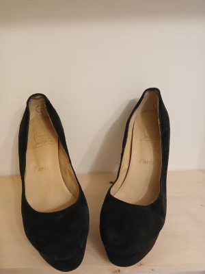 Svarta mocka pumps från Louboutin - Säljer ett par klassiska svarta pumps från Christian Louboutin i mocka med ikonisk röd sula. Skorna har hög klack och rund tå samt en rejäl plattform framtill. Perfekta för dig som vill ha en tidlös och lyxig look. Använda men har en del kvar att ge.