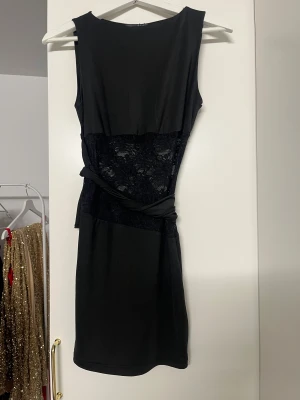 Svart kort klänning med spetsdetalj - Snygg svart klänning med spetsdetalj i midjan och omlottband. Ärmlös design och rund halsringning ger en stilren look. Perfekt för dig som gillar en enkel men ändå elegant stil. Materialet känns mjukt och följsamt mot kroppen.