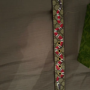 Gucci skärp med ormmönster - Säljer ett Gucci skärp med ikoniskt GG-mönster och en röd, vit och svart orm som slingrar sig längs hela bandet. Skärpet är gjort i beige canvas med detaljer i brunt läder och har guldfärgade metallhål. Perfekt accessoar för att lyfta din outfit.