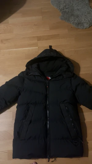 Svart pufferjacka zavetti canada - Svart pufferjacka från Canada Snow med huva och dragkedja framtill. Jackan har två stora fickor med dragkedja och är fodrad för extra värme. Perfekt för kalla dagar och har en clean, stilren look.