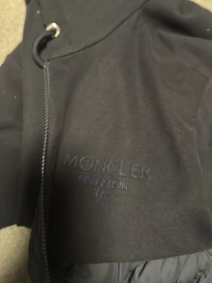 Svart Moncler pufferjacka M - Svart pufferjacka från Moncler med broderad logga på bröstet och klassisk Moncler-märke på ärmen. Jackan har dragkedja framtill och quiltad insida. Materialet är polyester och dun, perfekt för kyliga dagar. Snygg och stilren design med huva och justerbar snörning. Finns kvitto och pris lapp taggen funkar och moncler märket går och scanna!