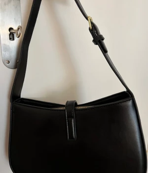 Svart axelväska från Saint Laurent - Snygg svart axelväska från Saint Laurent i slätt skinn med guldiga YSL-detaljer framtill. Väskan har en stilren, rektangulär form och justerbar axelrem med guldfärgat spänne. Dragkedja upptill och elegant design som verkligen sticker ut.